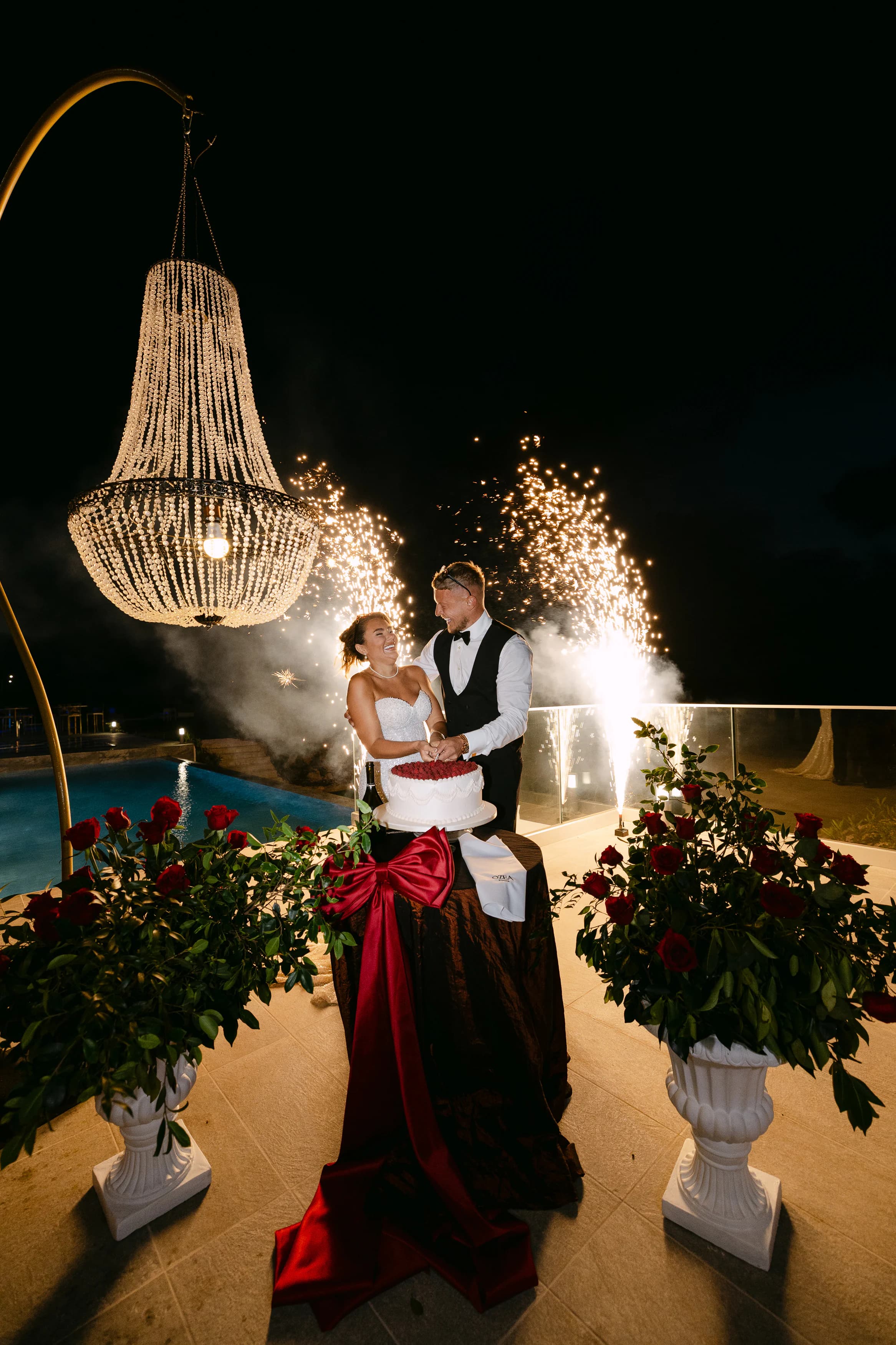 Bride and groom cutting the wedding cake at OZEA with sparkler fountains, crystal chandelier and red rose urns flanking them at night