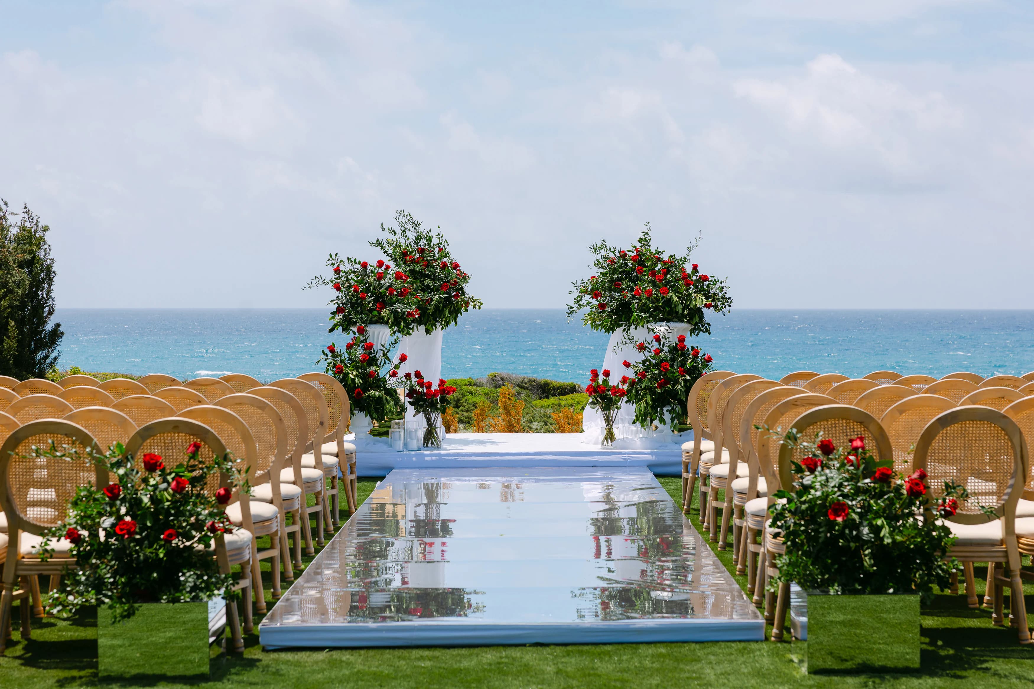 OZEA wedding ceremony aisle with mirrored runner, cane Louis-back chairs and red rose altar arrangements with the Mediterranean Sea behind