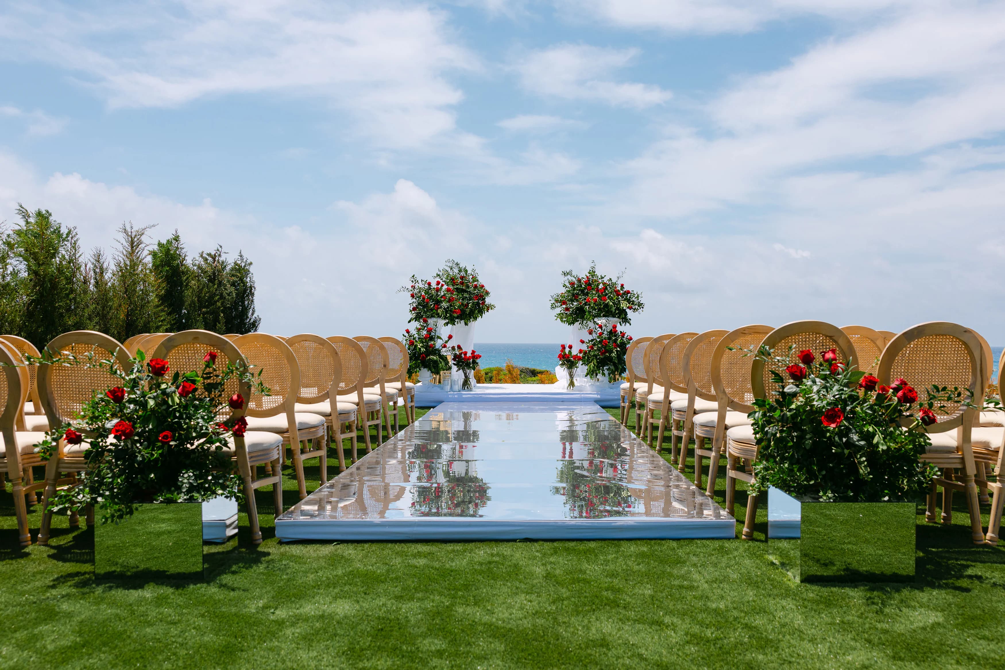 Wide view of the OZEA ceremony setup with cane chairs flanking a mirrored aisle and twin red rose pedestals against the sea