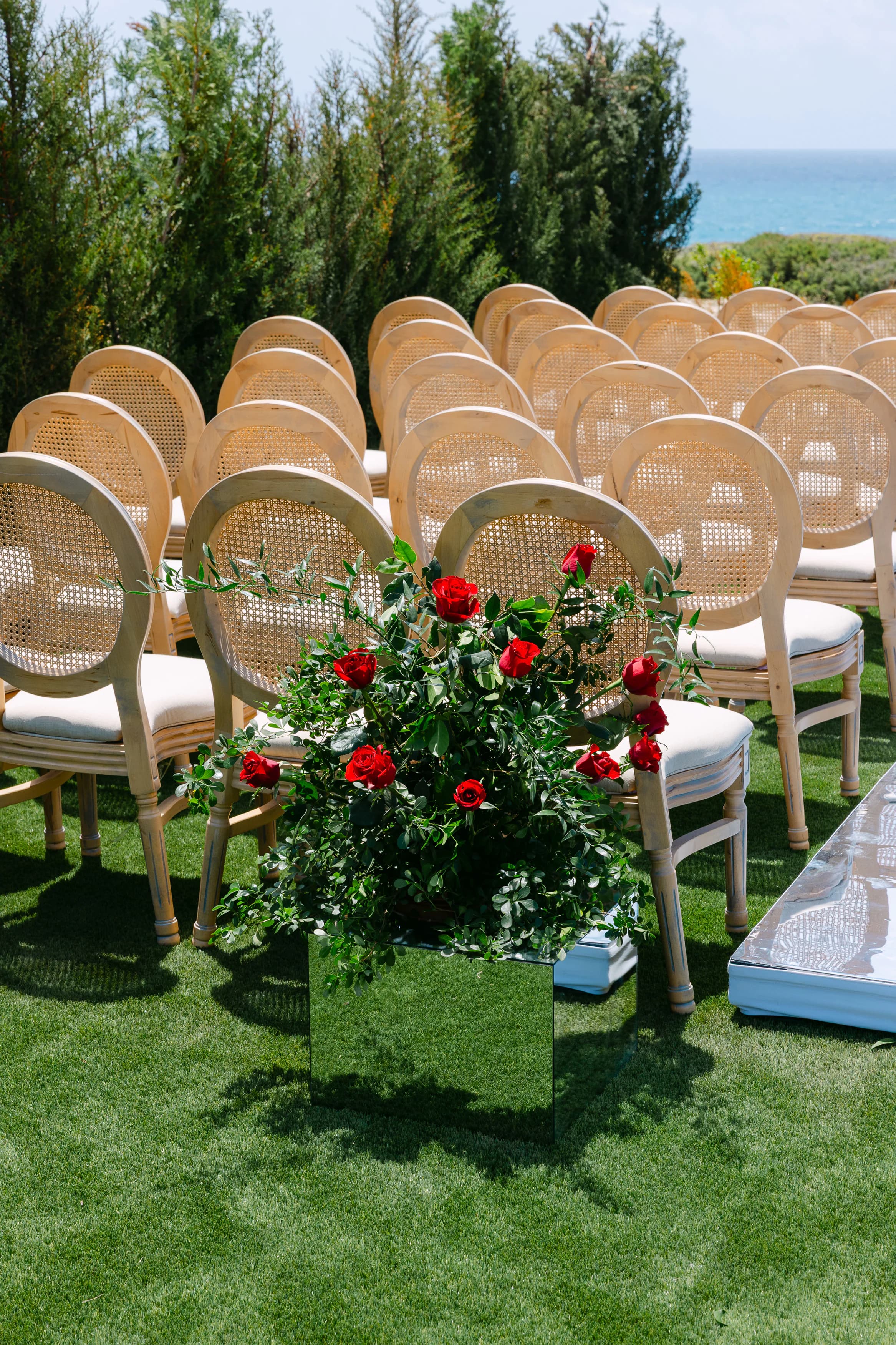 Mirrored cube aisle marker filled with red roses and trailing greenery beside cane chairs at the OZEA ceremony