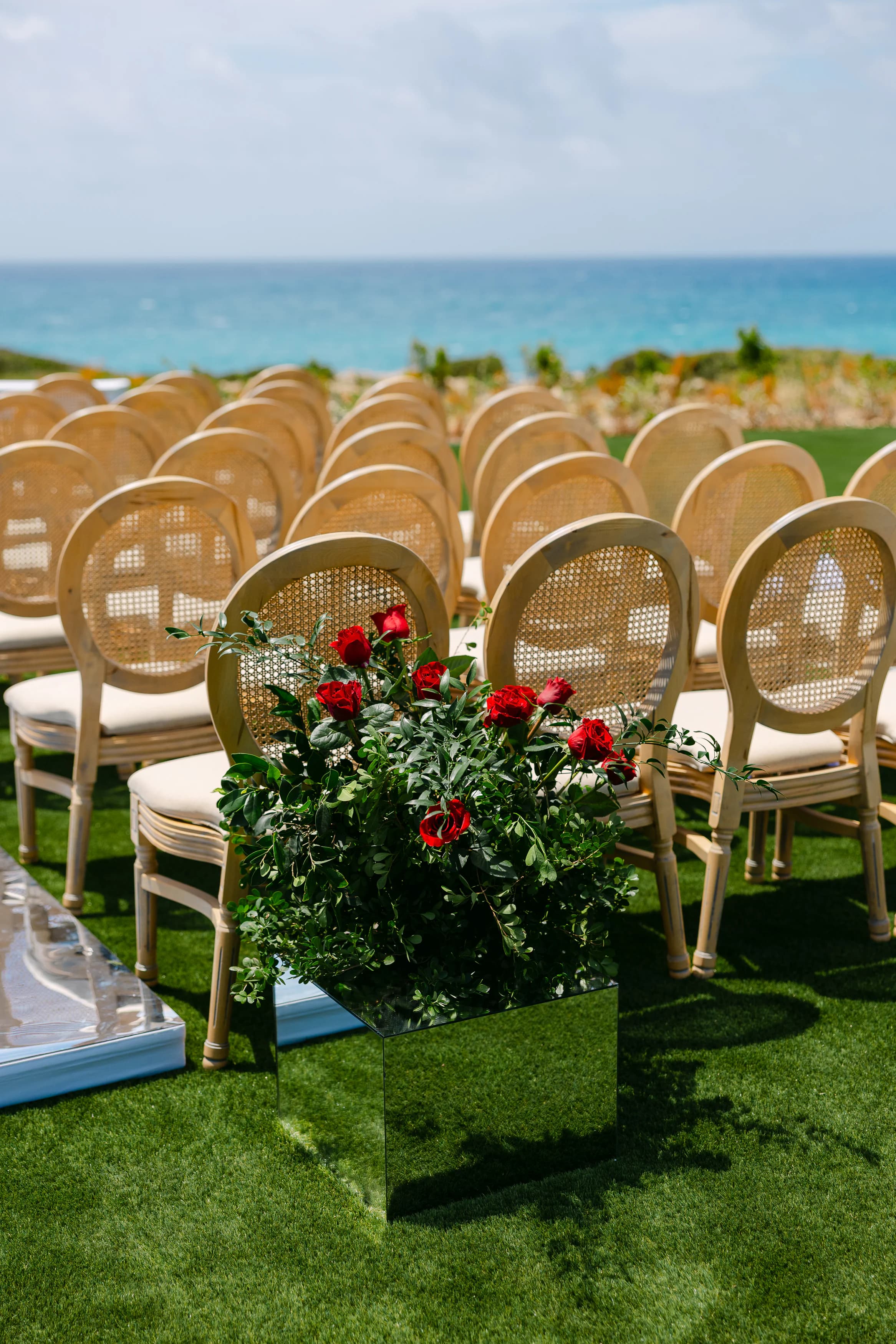Rows of cane Louis-style ceremony chairs at OZEA with a red rose aisle marker and the Sea Caves Paphos coastline in the background